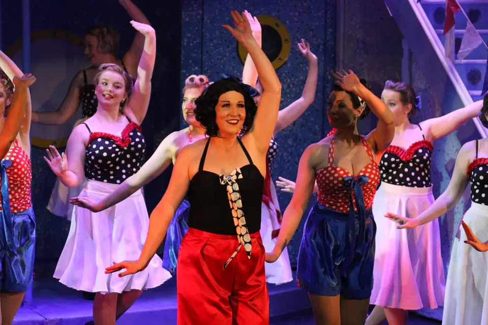 A performer in a black halter top and red trousers throws both arms up mid-number, the chorus around her in red, white and navy polka dots on a ship deck set.