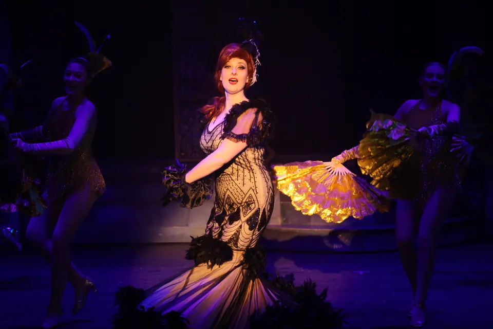 A performer in a beaded gown sings centre stage while dancers sweep gold feather fans either side of her, all under purple light.