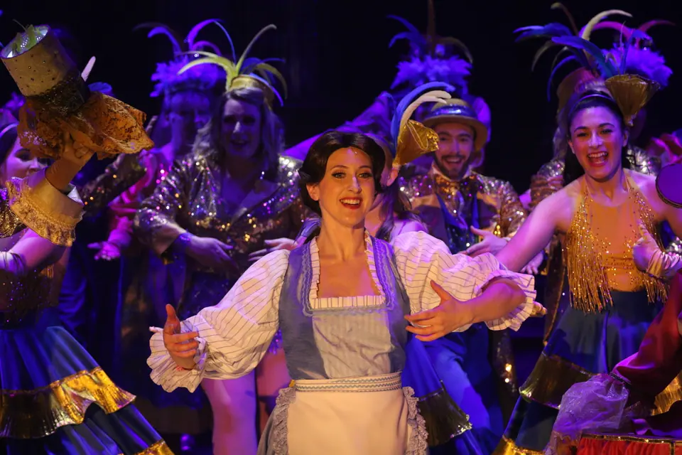 A performer in a blue bodice and white apron reaches wide, surrounded by dancers in sequins and tall feather headdresses under violet light.