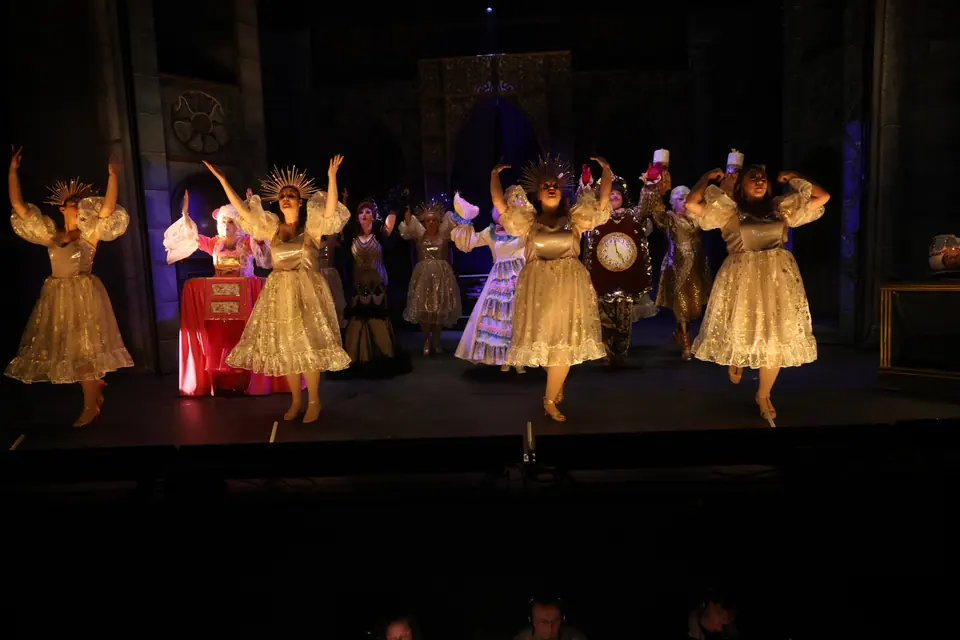 Performers in cream and gold dresses with sunburst headpieces raise their arms in a line, candle and clock costumes among them on a dark castle set.