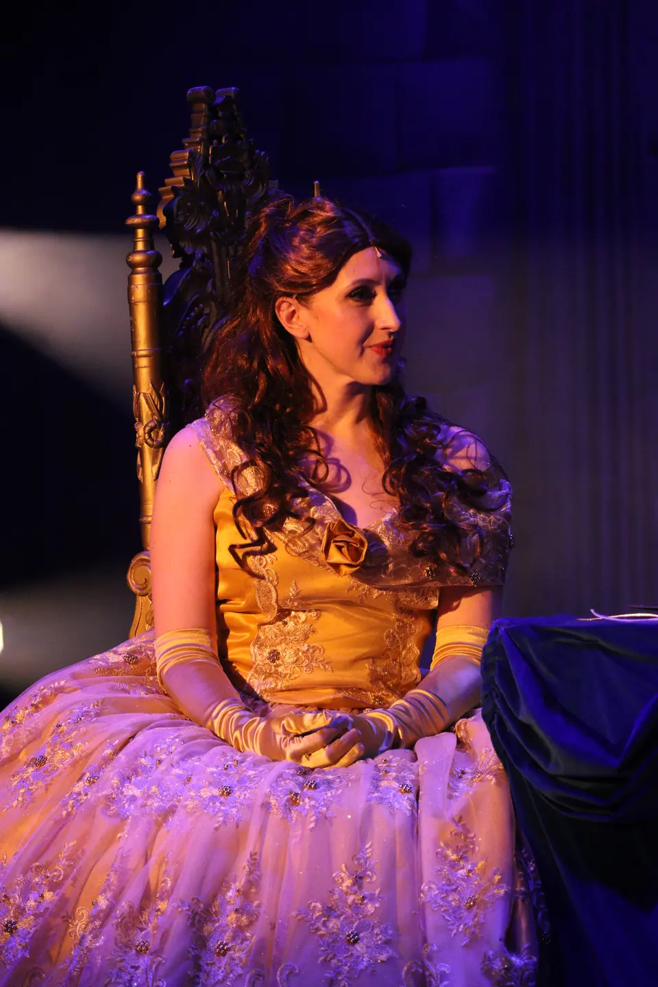 A performer in a gold ball gown sits on a carved gilt chair, hands folded, half in amber light and half in violet.