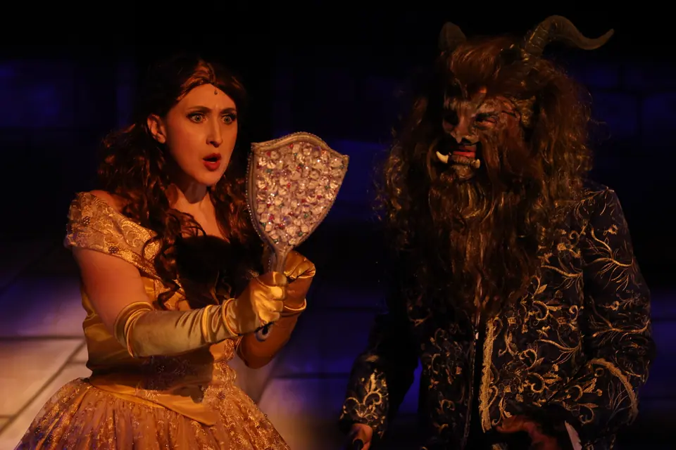 A performer in a gold gown holds up a jewelled hand mirror, mouth open, a figure in a beast mask and dark brocade coat beside her.