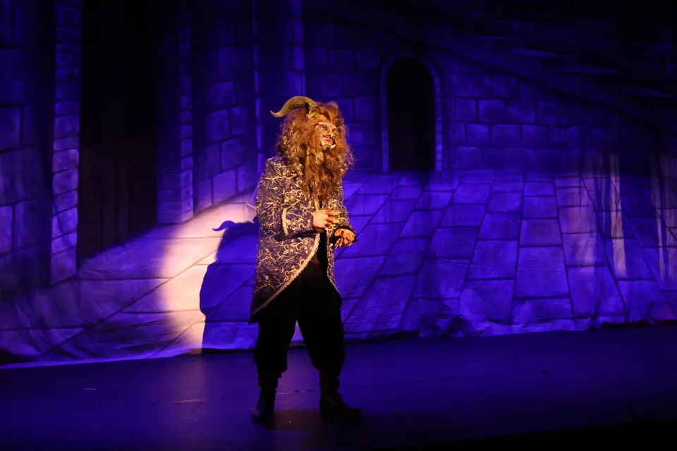 A lone figure in a beast mask and embroidered coat stands in a spotlight against a wide blue lit castle wall.