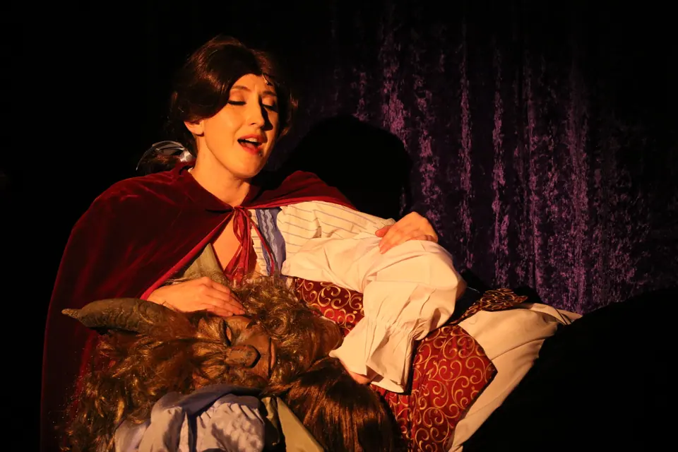 A performer in a red cloak cradles a figure in a beast mask across her lap, singing, against a dark purple drape.