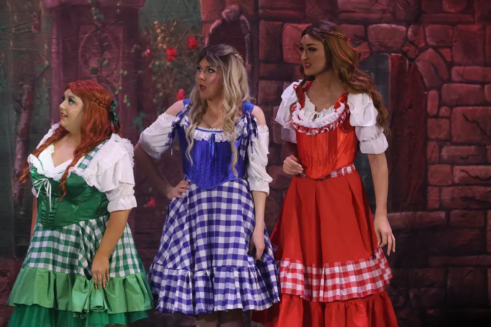 Three performers in green, blue and red gingham dresses stand together before a painted brick wall, all looking off to one side.