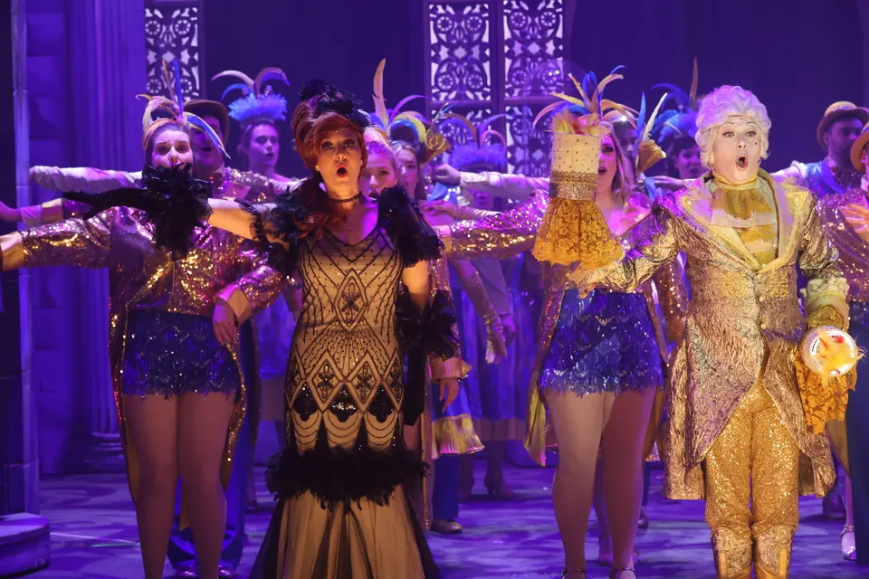 The company in sequins and feathered headdresses with arms flung wide, a performer in a beaded gown and another in gold livery leading the line.