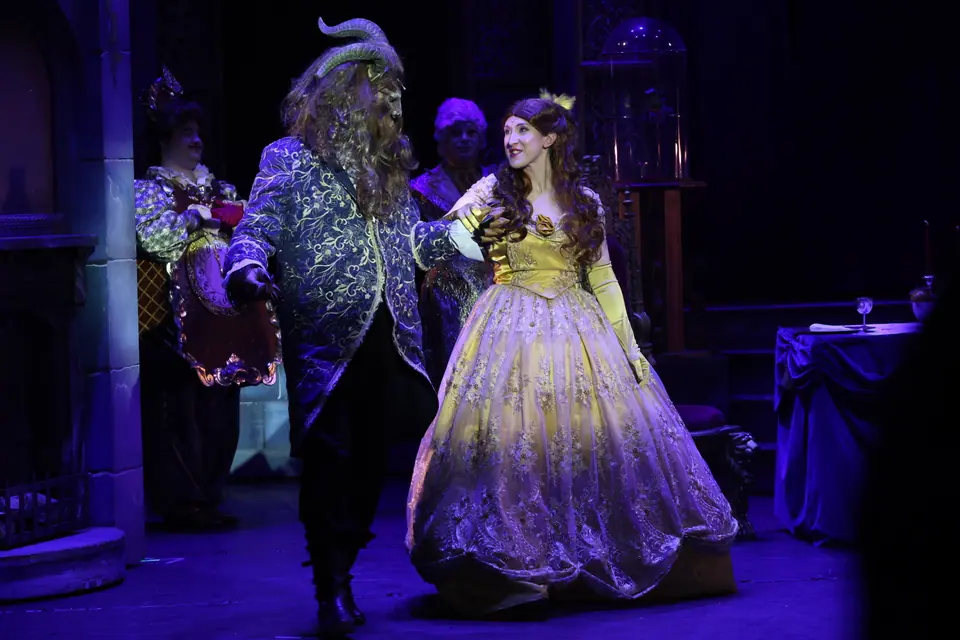 A figure in a beast mask and navy brocade coat leads a performer in a gold ball gown forward, the stage washed blue behind.