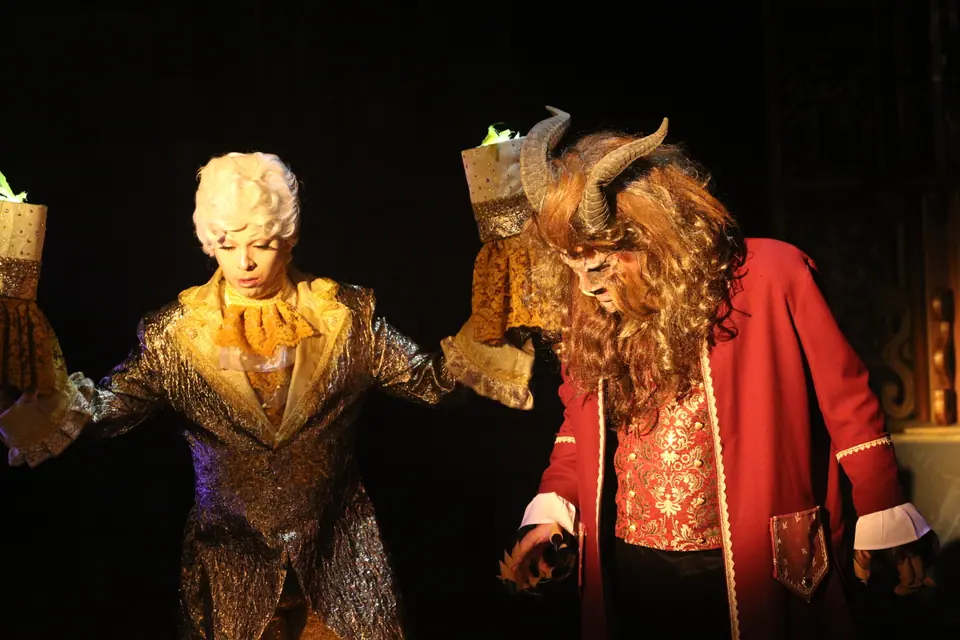 Two performers side by side in candlelight, one in a gold brocade coat and powdered wig, the other in a horned beast mask and red coat.