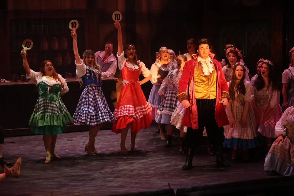 Three performers in bright gingham dresses hold up scorecards marked ten, a man in a red tailcoat downstage of a crowded tavern.