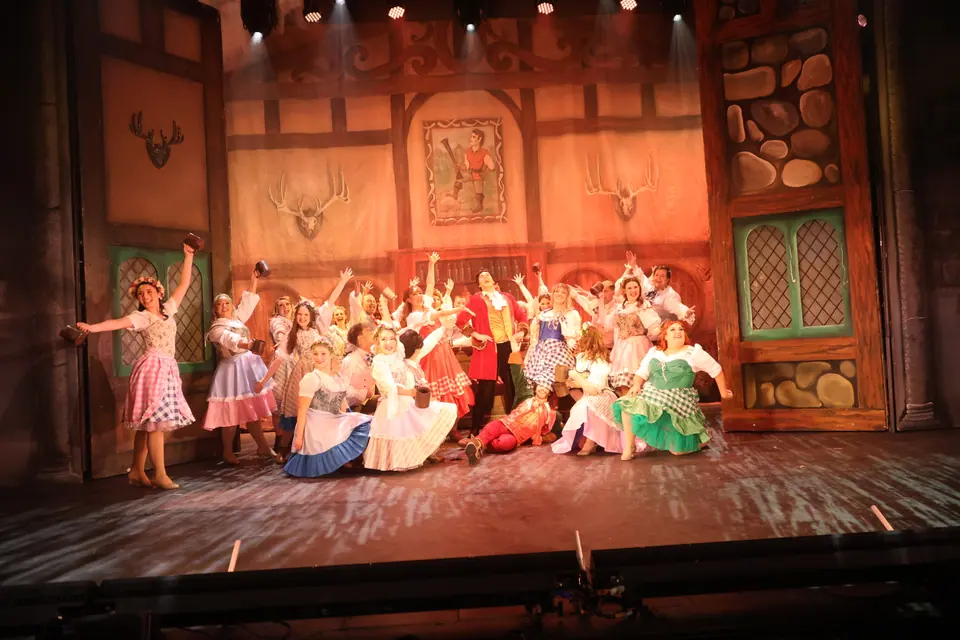 The full company across a tavern set with tankards raised, some kneeling at the front, the whole stage warm with amber light.