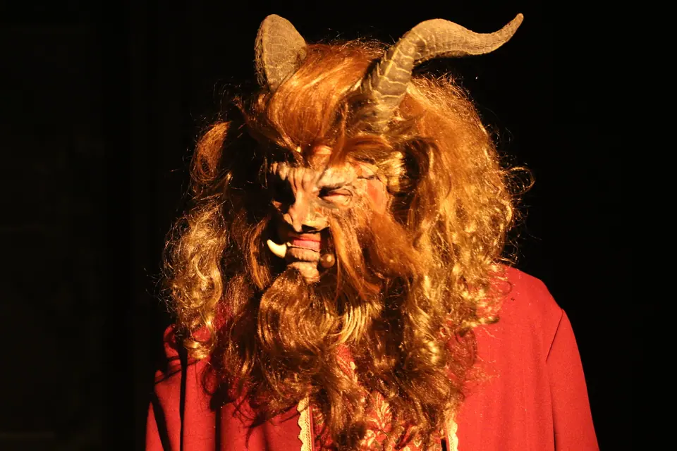 Close on a horned beast mask framed by a thick mane of tawny hair, red coat below, lit warm against black.