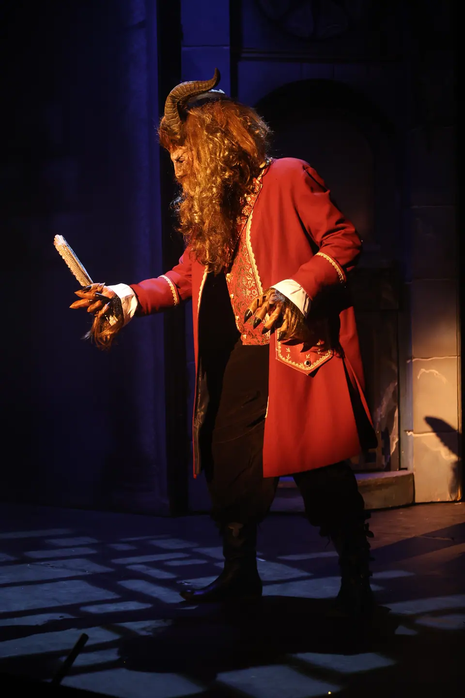 A performer in a horned beast mask and long red coat holds out a jewelled hand mirror on a blue lit stone set.