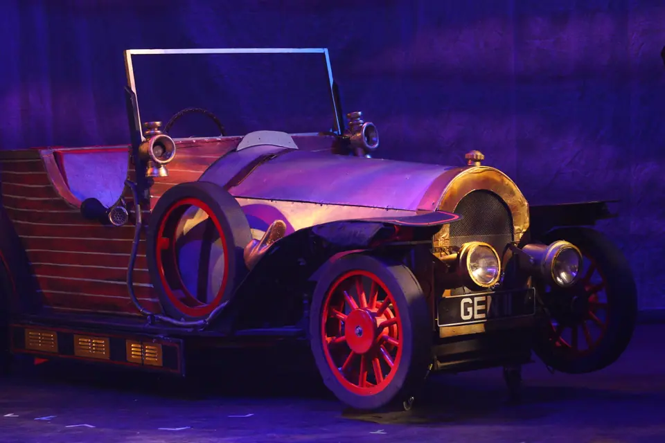 A brass and timber vintage motor car with red spoked wheels stands alone on a bare stage, lit deep purple from above.
