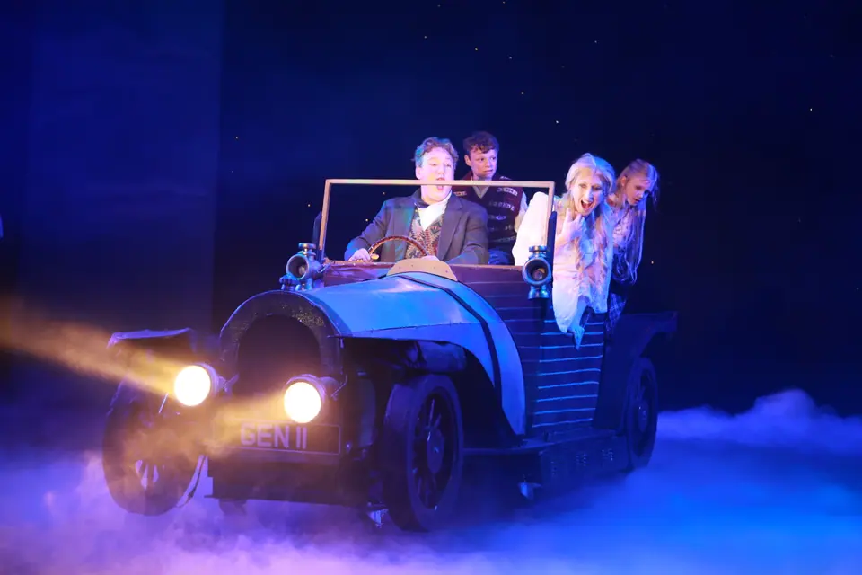 The vintage car faces the audience with headlamps blazing, four passengers leaning out, low fog rolling across a dark blue stage.