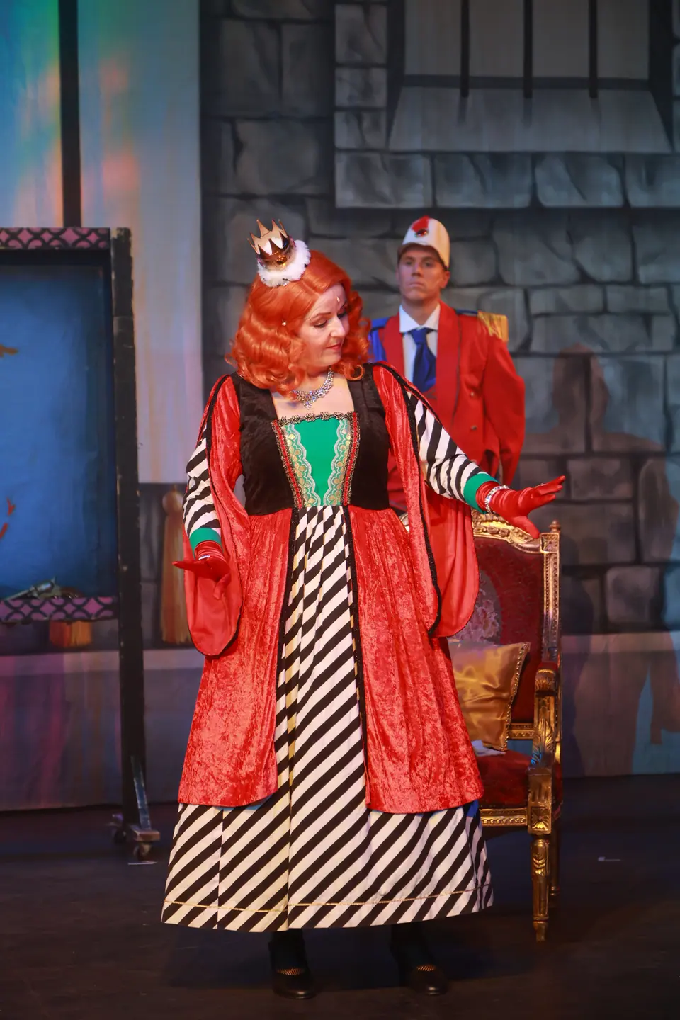 A performer in a small crown, red robe and black and white striped gown gestures beside a gilt throne, a guard in red standing behind.