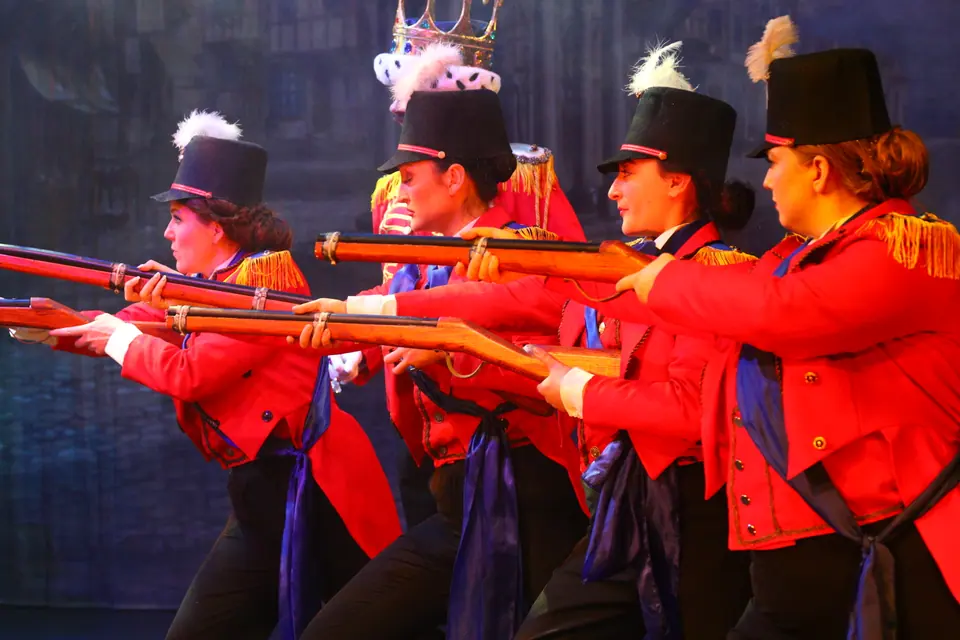 Four performers in red tunics and plumed black shakos level wooden rifles in a diagonal line, lit cold blue against painted stone.