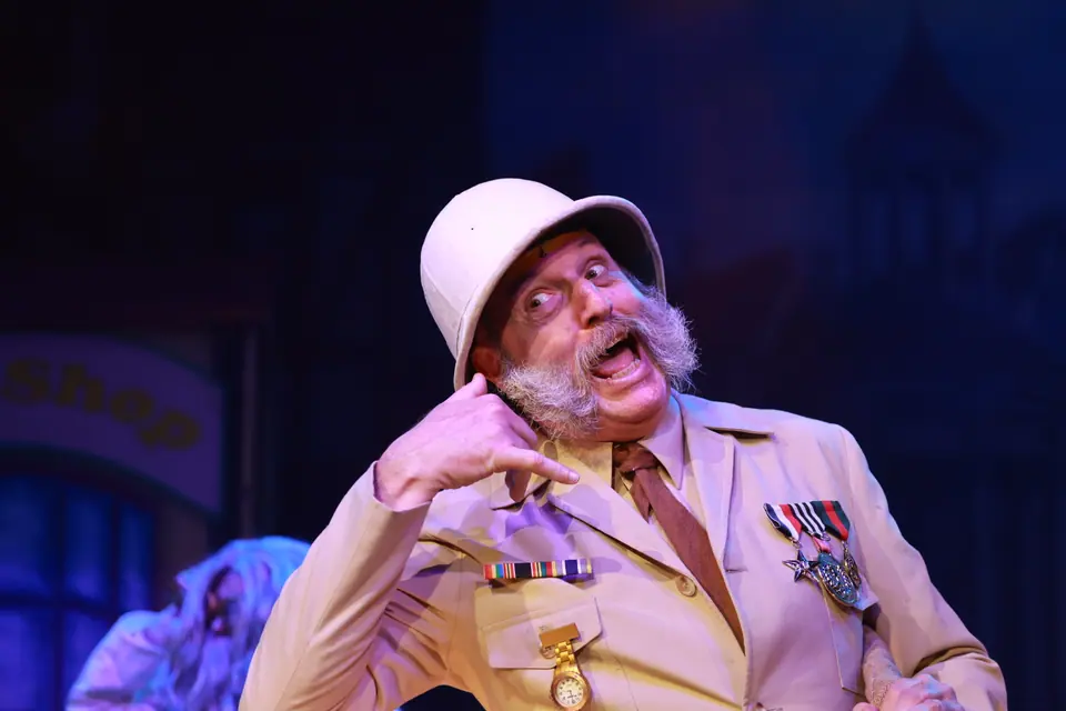 An older performer in a pith helmet and medalled khaki uniform pulls a face mid line, one hand raised beside his beard.