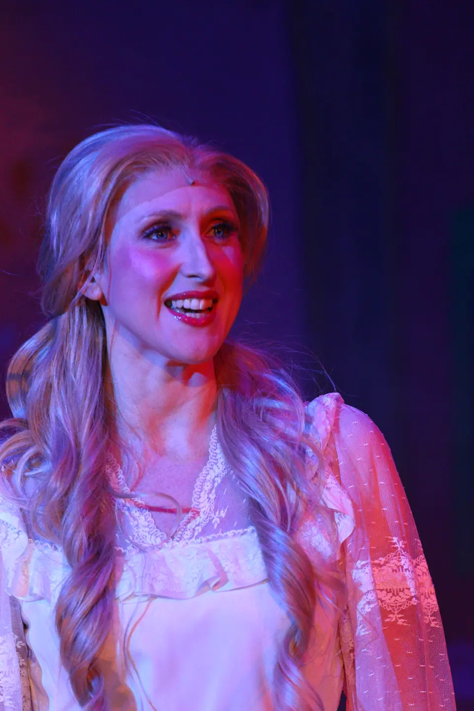 A performer with long fair hair and a white lace nightgown smiles off to one side, lit in pink and blue.