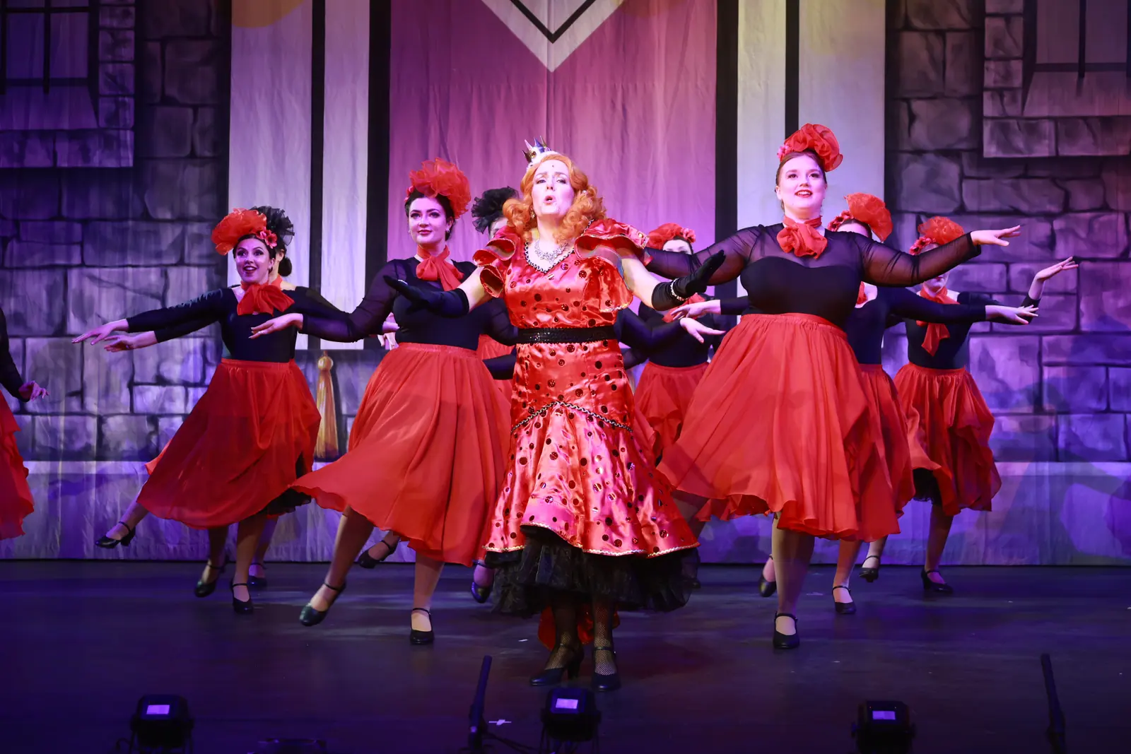 Dancers in black tops and full red skirts sweep their arms wide around a performer in a red sequinned gown and small crown.