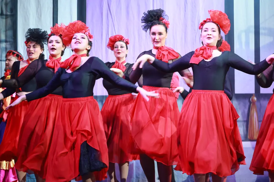 Six dancers in black tops, red skirts and red flowered headpieces move together in close view, skirts swinging out towards the camera.