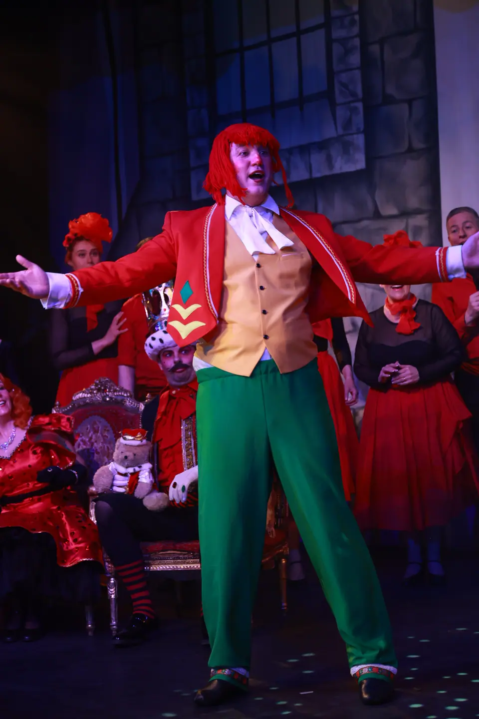 A performer in a red tailcoat, gold waistcoat, green trousers and a red yarn wig throws both arms out wide, the cast seated behind.