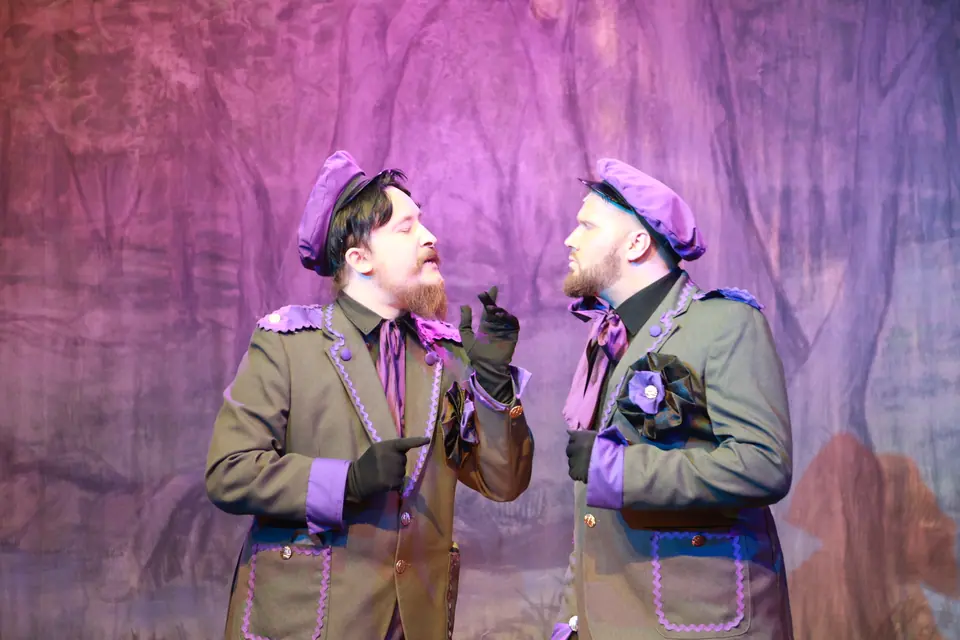 Two performers in olive greatcoats trimmed with purple and matching berets face each other mid argument against a painted purple forest.