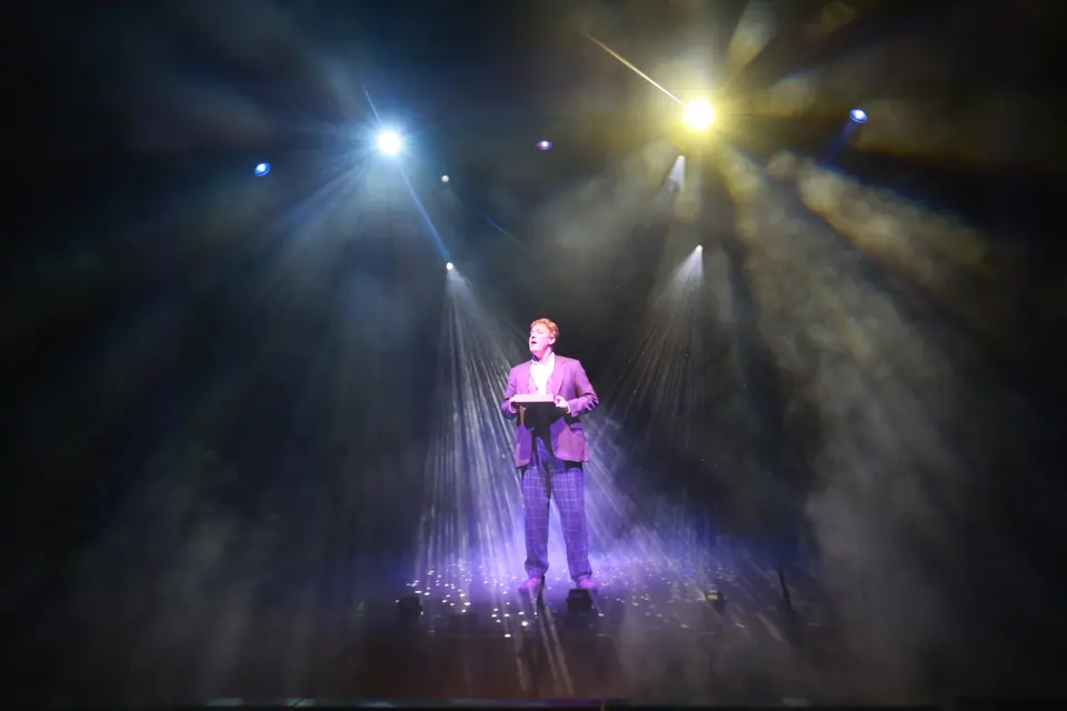 A lone performer in a mauve jacket and checked trousers sings in a narrow beam of light on a smoke filled stage.