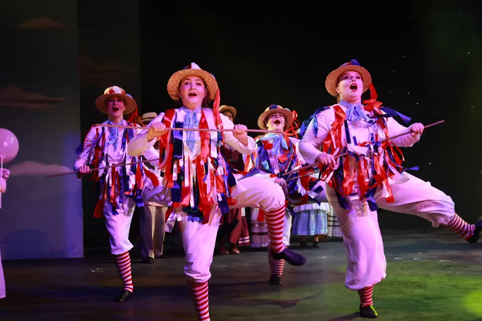 Performers in white costumes hung with red, white and blue ribbons and straw boaters kick high, poles held level across the line.