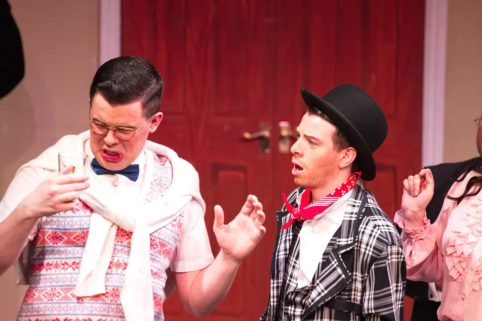 A man in a knitted tank top and bow tie pulls a face after sipping his drink while a man in a bowler hat and checked jacket reacts beside him.